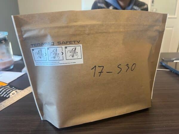 A brown paper safety bag labeled "TERA PAD SAFETY" with illustrations and safety icons. The handwritten text "17-S30" is on the bag. The bag is on a dark table, with a glass jar and paper visible in the background.