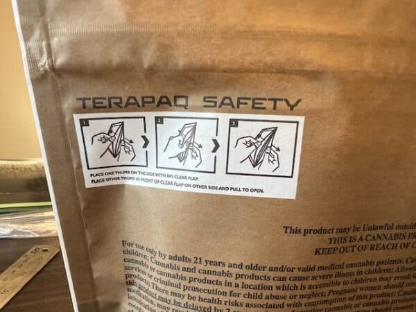 Close-up of a brown package with instructions for opening. The label titled "TERAPAQ SAFETY" shows three images: 1) Hold flap with thumb, 2) Place thumb in front of flap, 3) Pull to open. Small printed text is visible below the instructions.