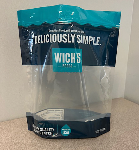 A transparent, empty food packaging bag, labeled "Wick's Foods," with text stating "Deliciously Simple." The bottom mentions "Premium Quality Always Fresh" and "Made in the USA." The bag is standing on a gray surface.