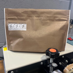 A brown paper bag sits on a machine with various buttons. The bag has a white label with safety instructions in small icons. The machine surface is light-colored, and the background is a gray wall.