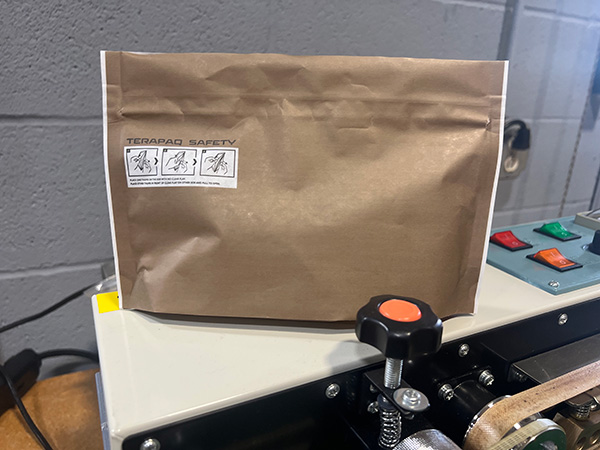 A brown paper bag sits on a machine with various buttons. The bag has a white label with safety instructions in small icons. The machine surface is light-colored, and the background is a gray wall.