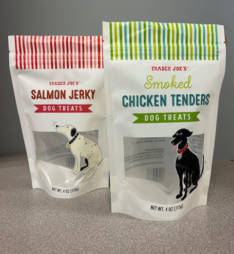 Two Trader Joe's dog treat bags stand on a surface. The left bag is labeled "Salmon Jerky Dog Treats" with a drawing of a white dog. The right bag is labeled "Smoked Chicken Tenders Dog Treats" with a drawing of a black dog.