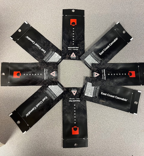 Eight black cannabis packaging bags arranged in a circular pattern on a gray surface. The bags have text and a red logo, forming a symmetrical star-like shape.